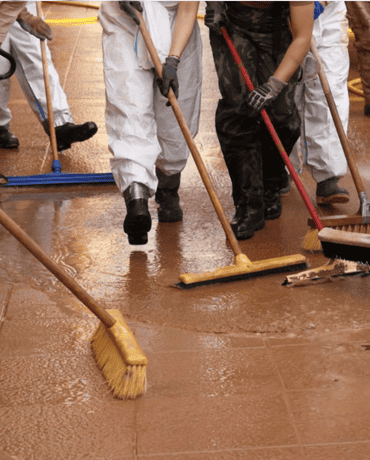 Several people in protective suits and gloves are using brooms to clean a wet floor.